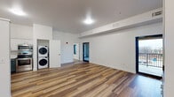 This single-floor unit in Bentonville, Arkansas offers a clean, contemporary design with an open-concept layout that maximizes usable space throughout. The main living area is expansive and bright, featuring warm wood-tone luxury vinyl plank flooring, crisp white walls, and flush ceiling lighting that gives the space a fresh, move-in-ready feel. Large black-framed doors provide direct access to an outdoor balcony, allowing natural light to filter into the living area and offering views of the surrounding neighborhood.

The kitchen is a standout feature, showcasing a two-tone cabinet design with white upper cabinetry and teal-blue lower cabinets paired with white countertops and a undermount sink. Stainless steel appliances, including a refrigerator, electric range, over-the-range microwave, and dishwasher, complete the well-appointed space. A dedicated nook adjacent to the kitchen houses a stacked washer and dryer unit, adding in-unit laundry convenience.

The bedrooms are carpeted for comfort and include closets with hanging rods. One bedroom features direct balcony access via a window and door combination. The two bathrooms mirror each other in design, each finished with large-format tile flooring, teal-green vanity cabinetry, white countertops, and well-lit mirrors, reflecting the cohesive color palette found throughout the unit.
