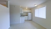 This single-floor space features an open layout that combines the kitchen and living area into one cohesive room. The kitchen is outfitted with white upper and lower cabinetry providing ample storage, a countertop with an integrated sink, and white appliances including a full-sized refrigerator and stovetop. The cabinetry and appliances share a clean, uniform white palette that contributes to a bright, tidy aesthetic. Light-toned tile flooring extends throughout the space, reflecting light and enhancing the sense of openness. The walls are finished in a neutral beige tone, complementing the overall understated color scheme. Two flush-mount ceiling light fixtures ensure consistent overhead illumination throughout the room. A large window fitted with vertical blinds along one wall allows natural light to fill the space, adding airiness to the interior. The open floor plan between the kitchen and the adjacent living area allows for flexible furniture arrangements. The overall ambiance is clean, minimal, and functional, making efficient use of the available space.
