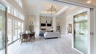 This elegantly designed single-floor community space offers a refined and welcoming atmosphere defined by its dramatic vaulted, shiplap-paneled ceilings and a coffered tray ceiling accent that frame a striking wrought-iron chandelier as a central focal point. The expansive main lounge area is finished with large-format, light-toned tile flooring that runs throughout, lending a clean, cohesive look to the open floor plan. A floor-to-ceiling mosaic tile accent wall serves as a bold architectural backdrop, flanked by dual flat-screen televisions and pendant lighting, alongside a built-in wet bar or beverage station — ideal for gatherings and events. The lounge is furnished with a comfortable seating arrangement, while an adjacent dining area accommodates a full table and chairs. A secondary sitting room offers a more intimate setting, featuring large windows that flood the space with natural light and views of lush surrounding greenery, accented by stylish pendant and wall sconce lighting, curated artwork, and a mix of upholstered accent chairs. Glass-paned French doors open directly to an outdoor deck, creating a seamless connection to the exterior. Throughout, the neutral color palette, layered lighting, and thoughtfully selected furnishings combine to create a polished, inviting environment.