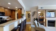 This single-floor residence at Apollo offers a well-designed layout across two bedrooms and two bathrooms, combining comfort with contemporary style. The open-concept living and dining area features warm hardwood-style flooring that extends throughout the main living spaces, creating a cohesive and inviting atmosphere.

A raised breakfast bar with granite or stone countertop detailing serves as a natural dividing point between the kitchen and living area, accompanied by sleek dark bar stools that add a modern touch. The kitchen features rich wood cabinetry, providing ample storage while contributing to the warm, sophisticated aesthetic of the home.

The living area is thoughtfully appointed with textured woven furnishings and accented by decorative metallic wall art, lending the space a curated, designer feel. A built-in media and shelving unit adds functional storage while maintaining the room's clean lines. Recessed lighting throughout ensures even, ambient illumination, complemented by accent lamps.

A wide hallway connects the living spaces to the bedrooms, lined with framed artwork and leading to the private quarters. Crown molding details along the ceilings add an elevated architectural finish throughout.