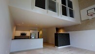 This two-story space makes an immediate impression with its dramatic double-height ceilings that flood the main level with an expansive, open sense of volume. The upper mezzanine level is framed by sleek, industrial-style metal railings and large windows that draw natural light deep into the interior, enhancing the airy atmosphere throughout.

On the main floor, a contemporary kitchen anchors one side of the open-plan layout, equipped with stainless steel appliances including a refrigerator and microwave, white cabinetry, and a clean white kitchen island or peninsula that provides both workspace and a visual divider. Light wood flooring runs throughout the main level, lending warmth and continuity to the space.

A distinctive row of dark, built-in lockers serves as a bold architectural element on the main floor, contrasting handsomely against an exposed brick accent wall behind them — a nod to industrial design sensibility. Recessed ceiling lighting provides even, ambient illumination across the open area.

The mezzanine level above offers additional functional space, accessible via the interior and overlooking the main floor below through the open railing system. Together, the two levels create a dynamic, loft-inspired environment that balances modern finishes with raw, textural details for a space that is both practical and visually compelling.