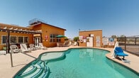 Located at 1330 New Harvest Pl in El Paso, Texas, this outdoor community amenity space centers around a generously sized freeform swimming pool with clear, blue-green water. The pool features a gradual entry point and reaches a depth of four feet, making it suitable for a range of swimmers. An ADA-compliant pool lift is installed poolside, ensuring accessibility for all residents. The expansive surrounding deck is finished in a light-colored concrete surface and is furnished with multiple lounge chairs and chaise lounges, ideal for relaxing under the El Paso sun. A shaded seating area with a green umbrella, table, and chairs provides a comfortable spot for outdoor dining or socializing. A dedicated pool house structure serves as a utility and equipment facility, with posted pool rules and safety signage prominently displayed. The entire pool area is enclosed by a wrought iron security fence, providing a safe and private environment. Adjacent to the pool, a children's playground equipped with a slide and climbing structure adds recreational value for younger residents. The warm earth-toned stucco exterior of the surrounding multi-story residential building complements the Southwest architectural aesthetic of the community, while mature trees add touches of greenery throughout the space.
