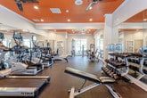 This single-floor fitness center is designed to support a comprehensive workout experience, combining striking aesthetics with a wide range of exercise equipment. The space is immediately distinguished by its bold orange ceiling, which contrasts sharply with the white walls and crown molding, creating an energetic and motivating atmosphere. Multiple ceiling fans are evenly distributed throughout, ensuring effective air circulation during workouts.

Natural light flows generously into the space through large windows that line the walls, offering views of surrounding greenery and keeping the environment open and inviting. Floor-to-ceiling mirrors are strategically positioned to visually expand the space and allow users to monitor their form during exercise.

The cardio zone is well-stocked with a row of treadmills and elliptical machines from True Fitness, alongside a Bodycraft rowing machine, catering to a variety of cardiovascular training preferences. The strength training area features a dedicated free weights section with a full rack of dumbbells, kettlebells, medicine balls, and a weight bench. Additionally, a selection of cable and resistance machines provides options for targeted muscle training.

The rubber flooring throughout provides durability and safety underfoot. A water cooler is conveniently available within the space. The overall layout is spacious and thoughtfully organized, allowing for ease of movement between equipment zones.