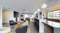 This single-floor space is a thoughtfully designed communal lounge that blends modern aesthetics with functional versatility. The open-plan layout creates a seamless flow between distinct zones, each serving a specific purpose while contributing to the overall inviting atmosphere.

A prominent bar counter with a crisp white quartz surface runs along one side of the space, flanked by warm wood-toned cabinetry and outfitted with leather-upholstered bar stools. A series of globe pendant lights hang overhead, casting a warm, ambient glow across the counter area.

The main seating area features an eclectic mix of contemporary furnishings, including leather lounge chairs and white accent chairs arranged around wooden side tables. A bold geometric patterned rug in yellow and grey anchors the seating zone, adding a vibrant design element against the large-format grey tile flooring.

Toward the back of the space, a projection screen is mounted for presentations or entertainment, accompanied by additional lounge seating. A reclaimed wood accent wall with an integrated television adds texture and warmth to this area.

Throughout the space, recessed lighting combined with large windows ensures ample illumination. The overall palette of whites, warm woods, and neutral tones creates a clean, sophisticated environment ideal for both professional and social use.