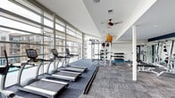This fitness center is defined by its striking floor-to-ceiling glass window wall, which runs along the length of the space and floods the interior with abundant natural light while offering open views of the surrounding building exterior. The single-level layout is thoughtfully organized to accommodate a range of workout activities. Along the windowed wall, a row of TRUE-brand treadmills provides a bright, energizing environment for cardio training. Toward the rear of the space, a multi-functional cable and power rack system serves as the centerpiece for strength and functional training. Adjacent to the rack, a well-stocked dumbbell rack holds a variety of free weights, while medicine balls and additional accessories are neatly organized on wall-mounted storage. A dedicated stretch and mat area occupies one corner, offering space for flexibility work and floor exercises. The ceiling features a ceiling fan that contributes to air circulation throughout the space. Neutral-toned carpet tiles cover the floor, and the white walls and ceiling enhance the overall brightness. The combination of ample equipment, natural light, and open layout creates a functional and inviting workout environment.
