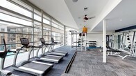 This fitness center is defined by its striking floor-to-ceiling glass window wall, which runs along the length of the space and floods the interior with abundant natural light while offering open views of the surrounding building exterior. The single-level layout is thoughtfully organized to accommodate a range of workout activities. Along the windowed wall, a row of TRUE-brand treadmills provides a bright, energizing environment for cardio training. Toward the rear of the space, a multi-functional cable and power rack system serves as the centerpiece for strength and functional training. Adjacent to the rack, a well-stocked dumbbell rack holds a variety of free weights, while medicine balls and additional accessories are neatly organized on wall-mounted storage. A dedicated stretch and mat area occupies one corner, offering space for flexibility work and floor exercises. The ceiling features a ceiling fan that contributes to air circulation throughout the space. Neutral-toned carpet tiles cover the floor, and the white walls and ceiling enhance the overall brightness. The combination of ample equipment, natural light, and open layout creates a functional and inviting workout environment.