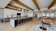 Located at 44 Braves Ave in Lawrenceville, GA, the Fieldhouse is a single-floor communal amenity space designed with a modern, sports-inspired aesthetic. The open-plan layout is anchored by exposed wooden ceiling beams and warm wood-look tile flooring that flow throughout, creating a cohesive and inviting atmosphere.

At the heart of the space is an expansive bar and kitchen area, featuring a long grey countertop lined with bar seating, dark wood cabinetry, a beverage cooler, and a subway tile accent wall with a mounted flat-screen TV. Distinctive yellow pendant lights hang overhead, adding a bold, stylish touch.

The lounge and social areas are thoughtfully arranged with high-top pedestal tables, mixed bar seating, and cozy leather lounge chairs. A full-size billiards table anchors one section of the space, while a comfortable seating nook near the entrance showcases the Fieldhouse branding on a feature wall.

A standout feature is the terrace-facing wall, equipped with large retractable glass garage-style doors that open to a window bench with colorful modular seating, offering direct views of the adjacent stadium and outdoor plaza. This connection to the outdoors floods the space with natural light and creates a vibrant, event-ready environment ideal for gatherings and community engagement.