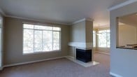 This single-floor residential space is thoughtfully designed with a focus on comfort and classic elegance. The living area serves as the centerpiece of the home, immediately drawing attention with its generous layout and warm, neutral color palette. Large, multi-pane windows line the walls, allowing abundant natural light to fill the room and offering pleasant views of the surrounding greenery outdoors.

A standout feature of the living space is the white-mantled fireplace with a sleek black firebox, positioned as a natural focal point within the room. The fireplace surround is finished in a clean, light-toned tile, adding a refined touch to the overall aesthetic. Crown molding runs along the ceiling perimeter throughout, lending an air of architectural sophistication and attention to detail.

The flooring is covered in neutral, plush carpeting that complements the warm beige tones of the walls, creating a cohesive and inviting atmosphere. An elegant pendant chandelier is visible in the adjoining area, hinting at a dining or secondary living space that benefits from the same natural light flooding through additional windows. The overall ambiance is clean, airy, and move-in ready, making it well-suited for a variety of interior design styles.