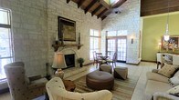 This single-story space showcases a warm, rustic aesthetic defined by floor-to-ceiling natural stone walls that lend a sense of solidity and timeless character throughout. The soaring ceiling is framed by rich, exposed wooden beams that draw the eye upward, emphasizing the dramatic vertical scale of the main living area. A ceiling fan is integrated seamlessly into the beam structure above.

The focal point of the main living area is a stone fireplace with a wooden mantel, framed by decorative accents and a piece of wall art above. The hearth is flanked by a classic table lamp and traditional fireplace tools, contributing to the cozy ambiance. Seating includes a cream upholstered armchair and a round leather ottoman, arranged over a striped area rug atop light-toned tile flooring.

Toward the rear of the space, a dining area with dark wood chairs and a small table sits adjacent to a set of elegant French doors. These doors flood the interior with abundant natural light and offer a view to the outdoors, creating a seamless connection between inside and out. The overall ambiance is inviting, rustic yet refined, blending natural materials with comfortable furnishings.