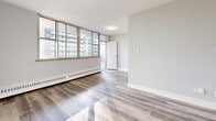 This 2-bedroom, 1-bathroom unit offers a clean, modern aesthetic across a thoughtfully arranged single-floor layout. Light wood-tone laminate flooring runs throughout, complementing the crisp white walls and trim that give the space a fresh, airy feel.

The entrance leads into a central hallway that connects all areas of the unit efficiently. The living room is a generous, sun-filled space featuring a wide bank of floor-to-ceiling windows that frame urban views and allow natural light to pour in. Baseboard heating runs along the perimeter, ensuring comfort year-round.

The kitchen is well-designed with white shaker-style cabinetry, granite-look countertops, a stainless steel sink, and white appliances including a stove and refrigerator. Overhead fluorescent lighting keeps the workspace bright and functional.

Both bedrooms are comfortably sized with neutral grey walls, light flooring, and windows overlooking the surrounding cityscape. The primary bedroom is notably spacious and includes a mirrored sliding-door closet. The second bedroom also benefits from natural light via a dedicated window with baseboard heating beneath.

The bathroom is clean and functional, featuring a full soaking tub with tiled surround and a sliding glass panel. Overall, the unit presents a bright, practical living environment with a cohesive, modern aesthetic.