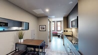 Located at 525 Main St in downtown Hartford, CT, this efficiently designed studio unit offers a thoughtfully organized single-floor layout comprising a living room, kitchen, bathroom, and closet. The open-concept design maximizes the use of space, with the kitchen and living/dining areas flowing seamlessly into one another. The kitchen is outfitted with sleek gray cabinetry, a built-in induction cooktop, and a textured tile backsplash, complemented by a wall-mounted microwave. A compact dining table with industrial-style chairs and stools provides a practical eating area. A pass-through window visually connects the kitchen to the dining space. The living and sleeping area features light-toned flooring, large windows with red-trimmed frames that invite natural light, and elevated ceilings that enhance the sense of openness. A sliding barn-style glass door separates the sleeping area, which includes a built-in wardrobe and wall-mounted TV. The bathroom is finished with floor-to-ceiling stone-textured tile, a soaking tub with shower, a sleek open-frame vanity, and warm tile flooring. Throughout, the unit maintains a modern, urban aesthetic with neutral tones and contemporary finishes.