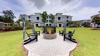 Located at 2222 Crow Ln in Myrtle Beach, South Carolina, this outdoor amenity space serves as a central gathering area within a well-maintained residential community. The focal point of the space is a circular stone fire pit, elegantly constructed with stacked masonry and surrounded by a decorative paver patio. Four black Adirondack chairs are symmetrically arranged around the fire pit, offering a comfortable and inviting setting for residents to gather and relax. The seating area is framed by lush, manicured green lawns that extend outward in multiple directions, providing an open and airy feel. Mature trees are thoughtfully planted within the grounds, adding natural shade and visual interest to the landscape. The backdrop features a multi-story residential building with a classic coastal aesthetic, finished in soft gray tones with white trim and covered balconies on each level — a design that is characteristic of the Myrtle Beach coastal region. The overall ambiance of this outdoor space is welcoming and community-oriented, blending functional amenities with attractive landscaping to create an environment well-suited for both quiet relaxation and social gatherings throughout the year.