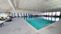 This indoor pool facility offers a well-appointed aquatic environment designed for year-round use. The centerpiece is a generously sized lap pool with crystal-clear turquoise water, framed by a wide surrounding deck that provides ample space for entry, exit, and poolside activity. The pool is equipped with lane markers, making it well-suited for structured swimming and exercise.

The facility is enclosed within a striking single-story structure featuring an exposed steel truss roof system that rises dramatically overhead, creating an airy, open atmosphere. The high, vaulted ceiling not only enhances the sense of spaciousness but also supports proper ventilation throughout the space. A continuous row of large, multi-pane windows lines one side of the facility, allowing natural light to filter in and offering a connection to the outdoors while maintaining a climate-controlled interior.

The walls are finished in a clean, neutral palette that complements the bright, reflective quality of the water. The overall design prioritizes functionality and comfort, making this indoor pool and cabana facility a versatile space suitable for lap swimming, recreational use, and aquatic programming throughout all seasons.