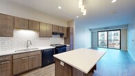 This single-floor unit showcases a thoughtfully designed open-concept layout with distinct living, sleeping, and cooking zones. The kitchen is a functional centerpiece, equipped with medium-toned wood cabinetry, a classic white subway tile backsplash, black appliances including a gas range, dishwasher, and refrigerator, and light-toned countertops. A kitchen island with a white surface and pendant lighting overhead adds both workspace and visual appeal. Wood-style flooring runs throughout the main living and kitchen areas, lending warmth and continuity.

The living area is bright and airy, benefiting from large floor-to-ceiling sliding glass doors that open to a private balcony with views of the surrounding building exterior, allowing abundant natural light to fill the space. The bedroom features plush carpeting, a large window with outdoor views, and direct access to a generously sized walk-in closet with wire shelving on multiple levels.

The bathroom is designed with accessibility in mind, featuring stainless steel grab bars, a wide-format vanity with an integrated sink, tile flooring, and a large mirror. The overall palette throughout is neutral and clean, creating a calm, move-in-ready ambiance.