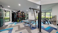 Located at 6316 Quail Hollow Rd in Charlotte, NC, this fitness center offers a thoughtfully designed single-floor space that accommodates a wide range of workout activities. The facility is organized across two interconnected areas, allowing for clear separation between cardio and strength training zones.

The main cardio area features soaring vaulted ceilings accentuated by bold blue accent stripes, creating a dynamic, energizing atmosphere. Treadmills, ellipticals, and a stationary bike are neatly arranged along large windows that invite natural light and provide views of the surrounding greenery. A full-length mirror at the end of the room enhances the sense of openness. Track lighting and recessed fixtures ensure even illumination throughout.

The adjoining strength and functional training area is well-stocked with cable machines, a weight bench, a dumbbell rack, TRX suspension trainers, resistance bands, kettlebells, and medicine balls. A large wall mirror amplifies the space visually. Posted fitness rules indicate an organized, community-oriented facility.

Throughout both areas, a striking chevron-patterned carpet in shades of blue, gray, and beige ties the design together cohesively. A sanitation station and private restroom access add convenience, making this a complete and practical fitness facility.