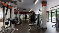 This modern fitness center is thoughtfully designed to accommodate a wide range of workout styles across a single, expansive floor. The space is unified by a bold color scheme of red, dark gray, and white accents that run along the upper walls and structural columns, giving the facility a dynamic, energetic atmosphere.

The main workout floor is divided into distinct functional zones. A strength training area houses a comprehensive selection of cable machines, multi-station weight systems, and adjustable benches, all set on durable rubber flooring. A dedicated free weights section features a full rack of dumbbells positioned in front of floor-to-ceiling mirrors, maximizing both space and visibility. Ceiling fans throughout help maintain airflow and comfort during intense sessions.

The cardio zone lines an entire glass wall, offering unobstructed views of lush, tree-lined greenery outside. Treadmills, ellipticals, and a rowing machine are arranged to take full advantage of this natural backdrop, with mounted flat-screen TVs providing additional entertainment.

A separate cycling studio offers a focused environment for spin sessions, equipped with multiple stationary bikes, individual monitors, and a central TV display, all set on wood-style flooring. Near the entrance, vending machines provide convenient post-workout refreshment alongside additional strength equipment. Natural light pours throughout the facility via large windows and glass doors, creating an open, inviting atmosphere.