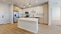 Located in Wesley Chapel, FL, this two-bedroom, two-bathroom home offers a thoughtfully designed layout with modern finishes throughout. The kitchen serves as the heart of the home, featuring a large white quartz island with pendant globe lighting, a herringbone-tiled backsplash, a combination of white and light wood cabinetry, and stainless steel appliances. Light wood-look flooring flows seamlessly through the main living areas, complemented by neutral wall tones that create a bright, airy ambiance. The living room benefits from abundant natural light, with large windows fitted with blinds and direct outdoor access through a glass door. Both bedrooms are carpeted and well-lit, each featuring windows that invite natural light and views of the surrounding greenery. The primary bedroom includes a dedicated walk-in closet with wire shelving for ample storage. The two bathrooms are finished with warm wood vanities, black hardware accents, white quartz countertops, and wood-look tile flooring. One bathroom features a soaking tub with marble-style tile surround, while the other provides convenient access to an additional walk-in closet. Each space reflects a clean, contemporary aesthetic designed for comfortable everyday living.