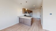This single-floor unit offers a thoughtfully designed open-concept layout that efficiently combines the kitchen and living area into one cohesive space. The kitchen is a standout feature, equipped with stainless steel appliances including an electric range, built-in microwave, and refrigerator. Rich walnut-toned cabinetry provides ample storage, while a peninsula with a granite-style countertop serves as a natural divider between the kitchen and living area. Pendant lighting above the peninsula adds a modern touch, complementing the recessed ceiling lights throughout.

Light wood-style flooring runs continuously across the entire unit, contributing to a clean, contemporary aesthetic. The living area benefits from a large window that invites generous natural light and offers outdoor views, with a wall-mounted HVAC unit providing climate control. A ceiling fan further enhances comfort in the space.

The bathroom includes a glass-enclosed shower and is positioned adjacent to a dedicated laundry area with stacked washer and dryer units, along with a closet featuring built-in shelving. White walls and bright, neutral tones throughout the unit create an open, airy ambiance, making the space feel larger than its compact footprint suggests.