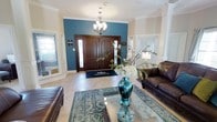 This single-story space makes a striking first impression through its thoughtfully designed entryway. The focal point of the entry is a bold teal accent wall that frames a stately set of rich, dark wood double doors, flanked by sidelights that invite natural light into the space. Crown molding runs along the ceiling perimeter, adding a refined architectural detail that elevates the overall aesthetic.

An elegant chandelier with classic candelabra-style lighting hangs above the entryway, casting a warm glow over the light-toned tile flooring below. A decorative area rug in complementary blue and neutral tones anchors the transition between the entry and the adjacent living area.

The living space is furnished with dark leather seating and centered around a glass-top coffee table, which displays a standout teal glass vase arrangement with white florals, adding a sophisticated decorative touch. Windows fitted with blinds on either side of the entry provide additional natural light while maintaining privacy.

The overall ambiance is polished and welcoming, combining classic architectural elements with contemporary decorative choices to create a cohesive and inviting interior.
