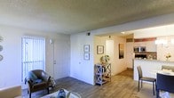 This single-floor residence is designed with an open-concept layout that promotes a sense of spaciousness and connectivity between living areas. The main living space features warm wood-style flooring that extends throughout, creating a cohesive and inviting atmosphere. A textured ceiling adds subtle visual interest overhead.

The entryway is framed by a classic white panel door, accompanied by a vertical window treatment that allows natural light to filter in. The adjacent wall provides a clean canvas, currently accented with framed artwork and a decorative X-frame console table, highlighting the versatility of the space.

The living area transitions naturally into the kitchen, which is visible through an open archway. The kitchen features light cabinetry, modern appliances including a built-in microwave, and a countertop that suggests ample prep space. The bright, well-lit kitchen contrasts warmly with the more subdued tones of the living area.

Overall, the space maintains a neutral color palette throughout, with white walls and warm flooring tones that create a welcoming, functional environment suited for everyday living.