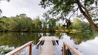 This single-level outdoor facility is thoughtfully designed to integrate with its natural wooded and waterfront surroundings. The centerpiece is a covered pavilion constructed with natural wood, featuring an open-air design supported by sturdy timber posts beneath a pitched roof with a wood-paneled ceiling. The pavilion accommodates seating with picnic tables and offers open sightlines into the surrounding forest canopy, making it well-suited for gatherings in a natural setting.

Connected to the pavilion is an extensive network of elevated walkways and boardwalks that wind through the wooded terrain. These pathways are framed by a combination of wood and metal cable railings, providing both safety and unobstructed views of the dense tree canopy and understory below. Interpretive signage and waste receptacles are positioned along the walkway, suggesting a public or educational use for the space.

A wooden dock extends from the shoreline out over a tranquil pond, flanked by cable railings and framed by mature trees draped with Spanish moss. The still water reflects the surrounding greenery, enhancing the peaceful, natural ambiance. The overall design prioritizes accessibility and immersion in the landscape, creating a seamless connection between built structures and the wooded, waterfront environment.
