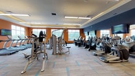 This modern fitness center is designed to support a comprehensive workout experience across a single, thoughtfully organized floor. The space is defined by bold orange and navy accent walls, tray ceilings with recessed and panel lighting, and patterned carpet tiles with teal accents that lend a clean, energetic atmosphere throughout.

The main exercise floor is generously proportioned, housing a robust selection of cardio equipment including treadmills, ellipticals, and stationary bikes arranged in rows. Multiple wall-mounted flat-screen televisions are positioned throughout for entertainment during workouts. Large windows along multiple walls invite abundant natural light and offer views of the surrounding outdoor area, including a glimpse of an adjacent pool.

A dedicated free weights zone features a full rack of dumbbells, medicine balls, and weight benches, complemented by floor-to-ceiling mirrors that maximize the sense of openness and support proper form. Shelving units provide organized storage for additional fitness accessories.

A separate studio room is accessible through a striking glass-panel wall with wood-framed double doors, offering a hardwood floor space ideal for group classes or individual training. Additional strength machines and functional training equipment are distributed throughout, rounding out the facility's extensive offerings.