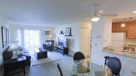 This single-floor residence is designed with an open-concept layout that seamlessly connects the living and dining areas, creating a cohesive and functional living environment. The living area is carpeted and anchored by a media console with a flat-screen television, complemented by a decorative floating shelf above. A patterned armchair and a low-profile coffee table rest atop a plush area rug, adding texture and comfort to the space. A sliding glass door along one wall allows natural light to filter in while providing direct access to an outdoor area, enhancing the overall sense of openness. A wall-mounted air conditioning unit sits beneath the window, ensuring year-round comfort. Overhead, a ceiling fan with an integrated light fixture serves both the living and dining zones. The dining area, positioned in the foreground, features a round glass-top table surrounded by dark chairs, lending a contemporary touch. Adjacent to the main living space, a hallway leads to additional rooms, with a glimpse of a secondary area visible beyond a partial wall. The overall palette is neutral and bright, with white walls and light-toned ceilings contributing to an airy, welcoming ambiance throughout.