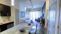 This single-floor residence showcases an open-concept design that seamlessly connects the dining and living areas, creating a cohesive and functional living space. The dining area features a marble-topped table beneath a contemporary pendant light, providing an elegant setting for meals. Hardwood-style flooring extends throughout, adding warmth and continuity to the layout.

The living room is a well-appointed space anchored by a ceiling fan and illuminated by natural light streaming through sliding glass doors that open to a private balcony. A bold navy accent wall adds a dramatic focal point, complementing the neutral tones of the surrounding décor. The room accommodates a comfortable seating arrangement centered around a round coffee table, with a patterned area rug defining the space.

Track lighting in the entryway area enhances visibility and adds a modern industrial touch to the overall design. The sliding glass doors at the far end of the living area provide outdoor views and allow ample natural light to brighten the interior. Overall, the space combines contemporary finishes with a thoughtfully designed layout, creating a comfortable and visually appealing living environment.