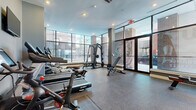 This fitness center offers a well-appointed, single-level space designed to accommodate a comprehensive workout experience. The room is finished with durable speckled rubber flooring throughout, providing both safety and functionality across all exercise zones.

Natural light is a defining feature of the space, with expansive floor-to-ceiling windows and glass-paneled entry doors lining one wall, offering open views of the surrounding streetscape and flooding the interior with daylight. Recessed lighting supplements the natural light, ensuring the space remains well-lit throughout.

The cardio zone is equipped with multiple Spirit treadmills, elliptical cross-trainers, and a Peloton spin bike, each positioned to take advantage of the exterior views. Wall-mounted flat-screen televisions are installed at convenient sightlines to enhance the cardio experience. The strength training area includes a multi-functional cable and weight machine, a dedicated free weight rack with dumbbells and medicine balls, and an adjustable weight bench. A rowing machine rounds out the equipment selection.

The neutral gray walls and clean architectural lines give the space a modern, professional aesthetic. Glass-paneled double doors provide entry access, contributing to the open and transparent character of the overall design.