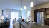 This single-floor residence showcases a thoughtfully designed open-concept layout that connects the kitchen, dining, and living areas into one cohesive space. The kitchen serves as a central focal point, anchored by a substantial island with a dark granite countertop that offers both prep space and casual seating for multiple chairs with nailhead-trim upholstery. Three elegant pendant lights with cylindrical shades hang above the island, providing focused task lighting while adding a refined visual element. A stainless steel undermount sink with a modern arc faucet is integrated into the island. Dark espresso cabinetry and a stainless steel refrigerator complete the kitchen's contemporary aesthetic. The open living area beyond benefits from generous natural light streaming through large windows fitted with vertical blinds, creating a bright and airy atmosphere. Neutral wall tones and light-colored flooring contribute to the space's clean, modern character. A glimpse into an adjacent room suggests a bedroom nearby, reinforcing the practical and connected flow of the floor plan. Overall, the space balances modern finishes with functional design.
