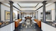 This single-floor community clubhouse is designed with a refined blend of classic and contemporary architectural elements throughout. The grand entrance lobby immediately sets an elegant tone, featuring gleaming dark tile flooring, soaring tray ceilings with bold dark crown molding, and stately decorative columns framing the space. Dual service counters with warm wood finishes line both sides of the lobby, offering a welcoming and organized reception area. Double glass entry doors at the far end draw in natural light and provide views to the exterior.

Beyond the lobby, the space opens into a spacious lounge area anchored by a chandelier, a fireplace, and a comfortable seating arrangement, with large windows dressed in sheer curtains that fill the room with soft, natural light. Lush potted plants add a fresh, inviting ambiance throughout.

The kitchen area is thoughtfully designed with gray cabinetry, bold black granite countertops, stainless steel appliances, and a striking patterned tile backsplash. A rustic wood-paneled breakfast bar with metal stools adds a casual, modern touch. An adjacent dining area features a round dark dining table with matching chairs, a ceiling fan, and direct access to the outdoors.

A dedicated fitness center offers multiple strength-training machines set against large windows with natural views. A quiet reading lounge with built-in cabinetry, warm hardwood flooring, and cozy seating rounds out this versatile and well-equipped facility.