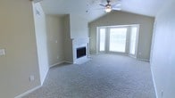 This single-floor home offers a thoughtfully designed living space highlighted by a standout great room that immediately captures attention. Vaulted ceilings rise overhead, creating an expansive sense of volume and openness, while a ceiling fan with a built-in light fixture provides both practical airflow and ambient illumination. The room is anchored by a classic fireplace framed in white-painted wood trim and clad in clean white tile, offering a timeless focal point suited to both relaxed evenings and entertaining. A large bay window adorns one wall, featuring multiple panes that invite generous amounts of natural light deep into the interior, further enhancing the airy atmosphere. The flooring throughout is wall-to-wall carpet in a neutral tone, adding warmth and comfort underfoot while complementing the soft, neutral wall colors. The overall palette is calm and versatile, providing an excellent canvas for a variety of interior styles. The combination of vaulted ceilings, abundant natural light, and the central fireplace gives this home a welcoming and comfortable ambiance that is both functional and inviting.