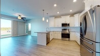 This well-appointed single-floor residence offers a thoughtfully designed open-concept layout that seamlessly connects the kitchen and living areas. The kitchen serves as the centerpiece, showcasing white shaker-style cabinetry with crown molding, granite countertops, and a classic white subway tile backsplash. A central island with a built-in sink and pendant lighting provides additional prep space and casual seating. Stainless steel appliances, including a French door refrigerator, electric range with microwave, and dishwasher, complete the modern kitchen ensemble. Light-toned hardwood flooring flows throughout the main living spaces, creating a bright and cohesive aesthetic. Recessed lighting and ceiling fans are installed throughout, ensuring consistent illumination. The bedrooms feature plush neutral carpeting and windows that allow natural light and exterior views. The primary bathroom includes an extended white vanity with granite countertops, a large mirror, and accessibility-conscious grab bars. A secondary full bathroom offers a tub-shower combination with clean white surrounds. The neutral gray wall tones and white trim work throughout the space lend a clean, contemporary feel, while sliding glass doors in the living area connect the interior to an outdoor space.