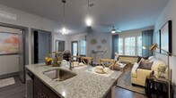 This single-floor residence offers a thoughtfully designed open-concept layout that seamlessly connects the kitchen, dining, and living areas. The kitchen is equipped with stainless steel appliances — including a refrigerator, electric range, and microwave — complemented by rich dark wood cabinetry and granite countertops. A kitchen island with an undermount sink and pendant lighting serves as both a functional workspace and a casual gathering spot. The dining area accommodates a round table and chairs, set atop hardwood-style flooring, with natural light filtering through blinds on a nearby window. The living room features a ceiling fan, large windows with drapery, and ample space for comfortable seating. Two bedrooms are carpeted and well-lit, each offering closet space and windows. The full bathroom includes a granite-topped vanity with dark wood cabinetry, a tiled tub-shower combination, and hardwood-style flooring. A dedicated in-unit laundry room houses a full-sized washer and dryer with overhead shelving. Neutral gray tones and consistent finishes throughout the space create a cohesive, modern aesthetic well-suited for everyday living.