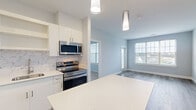 This single-floor residence in East Providence, Rhode Island, offers a thoughtfully designed layout with modern finishes throughout. The kitchen serves as a central highlight, featuring crisp white cabinetry, a marble-style tile backsplash, quartz countertops, and a full suite of stainless steel appliances including a range, microwave, refrigerator, and dishwasher. A contrasting-base island with a white quartz top provides additional prep space and casual seating, illuminated by sleek pendant lighting above. Light wood-tone luxury vinyl plank flooring extends through the kitchen and main living areas, lending a clean, contemporary feel.

The living area is generously proportioned, bathed in natural light through large windows that frame views of the surrounding neighborhood. Bedrooms are finished with plush neutral carpet and feature windows that bring in additional daylight. Closet space includes a walk-in with wire shelving for organized storage. The bathroom is appointed with wood-look tile flooring and includes a full soaking tub with shower. Soft gray-blue walls throughout the unit create a calm, cohesive palette that complements the modern finishes.
