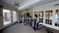 This single-level fitness space is thoughtfully designed to support an effective workout routine in a refined setting. The room is outfitted with quality cardio equipment, including treadmills and an elliptical trainer, arranged to allow comfortable movement between machines. A large floor-to-ceiling mirror along one wall visually expands the space while allowing users to monitor their form during exercise. The neutral taupe walls and carpeted flooring contribute a clean, polished aesthetic that feels more upscale than a typical gym environment. Natural light enters through multiple windows, keeping the space bright and inviting. Elegant white-framed French doors add a distinctive architectural touch, connecting the fitness area to an adjacent space and enhancing the sense of openness. Ceiling fans are installed to ensure proper air circulation during workouts. Crown molding along the upper walls adds a refined finishing detail consistent with the overall design sensibility of the space. The combination of functional equipment, natural light, and thoughtful design elements makes this fitness room a practical and pleasant environment for daily exercise.