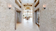 This expansive single-floor amenity space offers a sophisticated and welcoming atmosphere designed for comfort and social gathering. The entry corridor makes an immediate impression with its travertine stone-clad walls, large-format tile flooring, and soaring ceilings that draw natural light deep into the interior. A prominent arched window above the main entrance frames an outdoor view, enhancing the sense of arrival.

Beyond the entry, the space opens into a generously proportioned lounge area anchored by a floor-to-ceiling stacked stone fireplace with a mounted television above the mantle. The lounge features a warm wood-paneled coffered ceiling, pendant lighting, and an open layout furnished with neutral, contemporary seating arrangements.

Adjacent to the lounge, a sun-drenched gathering area is defined by dramatic floor-to-ceiling glass curtain walls that offer panoramic views of an outdoor pool and surrounding palm-lined landscape, seamlessly blending interior and exterior spaces.

A separate wing, accessible through wood-framed French doors with glass panels, provides a more private area suited for meetings or focused activities. Throughout, large-format tile flooring, warm wood trim accents, and carefully curated furnishings create a cohesive, resort-inspired aesthetic.