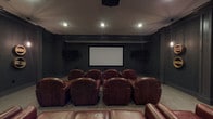 Located at 4125 Hayden Lofts Pl in Dublin, Ohio, this dedicated home theater offers a true cinematic experience within a single-floor layout. The screening room is designed with immersive entertainment in mind, centered around a large fixed projection screen mounted on a dark-painted accent wall, paired with a ceiling-mounted projector for crisp, high-quality image delivery. Recessed lighting is strategically placed in the ceiling to provide ambient illumination without interfering with the viewing experience. Flanking the main screen are wall-mounted displays, adding versatility to the room's entertainment capabilities. The seating arrangement consists of multiple rows of rich, brown leather club chairs with wide armrests and deep cushioning, providing both comfort and a classic theater aesthetic. A spacious leather sofa is also present at the front of the room, accommodating additional viewers. The room's dark walls and neutral carpeted flooring contribute to sound absorption and an enveloping, cinema-like atmosphere. The overall design prioritizes function and comfort, making this theater an ideal space for film screenings, sports viewing, or multimedia presentations.