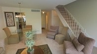 This three-floor home offers a well-organized layout designed for comfortable everyday living. The main level centers around a spacious living area finished with neutral carpet flooring and light-toned walls that create a clean, airy atmosphere. A classic white-railed staircase with carpeted treads serves as a prominent architectural feature, rising from the main living space and connecting the floors with a timeless aesthetic.

The living area accommodates a comfortable seating arrangement, including an armchair and ottoman, and flows naturally toward a glass-topped coffee table and additional seating. A decorative indoor plant adds a touch of warmth and greenery to the space. Recessed and ceiling-mounted lighting provides consistent illumination throughout.

Adjacent to the living area, a glimpse into the dining space reveals bar-height seating and a transition into what appears to be a kitchen beyond. A hallway extends toward the rear of the main level, leading to additional rooms and functional spaces. The open sightlines between areas enhance the sense of spaciousness across the floor. With three levels of living space, this home offers ample room for a variety of uses and configurations.