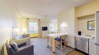 This thoughtfully designed suite offers a seamless blend of comfort and functionality across a single-floor layout. The open-concept living and sleeping area is bathed in natural light from multiple large windows dressed with soft yellow drapes, creating a warm, inviting atmosphere. The living space features plush gray seating — including a sofa and armchair — a flat-screen TV atop a white dresser, and a full-length mirror near the entry. A high-top dining table with bar-style seating provides a dedicated space for meals or work. The sleeping area flows naturally from the living space and includes a comfortable queen bed with wall-mounted lighting. The fully equipped kitchen is outfitted with stainless steel appliances, including a full-size refrigerator, dishwasher, and microwave, alongside white cabinetry and ample counter space — ideal for preparing meals in-suite. The bathroom features a split layout, with a vanity area separated from the water closet, which is accented with light blue walls and tile flooring. Throughout the suite, soft yellow and neutral tones, striped carpeting, and ceiling fans contribute to a relaxed, residential ambiance well-suited for extended stays.