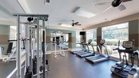 This fitness center offers a bright, well-appointed space designed to support a comprehensive workout experience. The room is lined with large windows that invite abundant natural light and provide views of lush greenery outside, creating an energizing atmosphere for exercise. A skylight further enhances the sense of openness, while a ceiling fan ensures comfortable airflow throughout.

The cardio area features a row of treadmills and elliptical machines positioned along the windowed wall, allowing users to enjoy the outdoor scenery during their workouts. A mounted flat-screen television adds entertainment options for those longer sessions. On the opposite side, a multi-station cable machine provides versatile strength training capabilities, complemented by additional free-standing workout equipment.

Full-length mirrors span one wall, visually expanding the space and allowing users to monitor their form. The flooring is durable and fitness-appropriate, designed to withstand heavy use. The walls are finished in a tasteful two-tone color scheme with soft blue-gray and neutral banded accents, accented by crisp white crown molding and a textured ceiling. The overall ambiance is clean, functional, and welcoming, making this fitness center a practical and pleasant amenity for daily use.