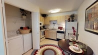 This single-floor space is thoughtfully designed to maximize functionality within a compact layout. The kitchen serves as a central feature, outfitted with light wood cabinetry that extends to the upper walls, offering generous storage throughout. A full suite of appliances is included, featuring a white refrigerator, electric range, built-in microwave, and dishwasher. The sink is positioned beneath upper cabinets along the back wall, and the countertops provide adequate workspace for meal preparation. Overhead, a flush-mount ceiling light provides bright, even illumination across the kitchen area. A notable practical feature is the in-unit laundry closet, positioned just off the kitchen, housing a full-size washer and dryer behind bi-fold doors, with shelving above for additional storage. Adjacent to the kitchen, the dining area accommodates a round dark-wood table with seating, anchored by a bold circular patterned area rug in red, cream, and green tones. Decorative touches throughout, including potted plants atop the cabinets and floral accents, contribute to a warm, lived-in ambiance.