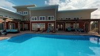 This outdoor amenity area centers around a large, rectangular swimming pool with vivid blue water, serving as the focal point of the space. The pool is surrounded by a spacious deck finished in neutral pavement, furnished with multiple reclining lounge chairs positioned to take advantage of open sky and natural light throughout the day. A safety ring is mounted nearby, reflecting attentive facility management.

The building backdrop features a combination of red brick along the lower level and neutral-toned lap siding on the upper story, giving the structure a classic yet approachable character. Large sliding glass doors and French doors provide direct access between the indoor spaces and the pool deck, facilitating seamless indoor-outdoor flow. A covered pergola-style overhang extends along part of the building facade, offering a shaded outdoor seating area furnished with dark wicker furniture and teal cushions — ideal for relaxing without direct sun exposure.

Multiple large windows along both levels allow natural light to filter into the interior while offering views of the pool area. The overall ambiance is that of a well-maintained communal outdoor retreat, designed to accommodate both active recreation and leisurely relaxation in an open-air setting.