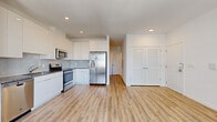 This thoughtfully designed 1-bedroom, 2-bathroom unit offers a clean, contemporary aesthetic throughout its efficiently utilized single-floor layout. The open-concept kitchen and living area serves as the heart of the space, featuring crisp white gloss cabinetry that extends to the ceiling for maximum storage, granite countertops, a subway tile backsplash, and a full suite of stainless steel appliances including a range, over-the-range microwave, dishwasher, and a side-by-side refrigerator with an in-door water and ice dispenser. Light wood-look plank flooring runs continuously throughout, lending warmth and visual cohesion to the space. The living area is bathed in natural light through large double windows that frame open exterior views, complemented by recessed lighting overhead. The bedroom offers a comfortable, well-proportioned retreat with the same wood-look flooring and easy access to a closet. The two bathrooms are finished with large-format white marble-style tile flooring, modern gray vanity cabinetry, white countertops, and mirrored medicine cabinets. A dedicated utility area with an electrical panel is also present. The overall ambiance is bright, clean, and move-in ready, reflecting a modern and functional design sensibility.