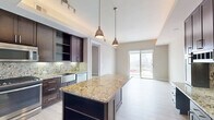 Located at 2212 McKinney Ave in the heart of Dallas, TX, this one-bedroom, two-bathroom unit offers a thoughtfully designed single-floor layout with modern finishes throughout. The gourmet kitchen is a standout feature, showcasing rich dark wood cabinetry, striking granite countertops with a decorative backsplash, stainless steel appliances including a French door refrigerator, gas range, and microwave, and a large central island with pendant lighting. A tray ceiling with ambient lighting adds architectural depth to the kitchen space. Light wood-tone flooring runs throughout, creating a clean and cohesive aesthetic. The open living area is filled with natural light from expansive floor-to-ceiling sliding glass doors that lead to a private balcony with sweeping Dallas city skyline views. The bedroom features neutral tones, hardwood-style flooring, and a window with additional city views. The primary bathroom is elegantly appointed with dark wood vanity cabinetry, granite countertops, large-format tile flooring, and a frameless glass-enclosed walk-in shower. A second bathroom provides additional convenience. In-unit laundry with a stacked washer and dryer rounds out the practical amenities of this well-appointed urban residence.
