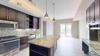Located at 2212 McKinney Ave in the heart of Dallas, TX, this one-bedroom, two-bathroom unit offers a thoughtfully designed single-floor layout with modern finishes throughout. The gourmet kitchen is a standout feature, showcasing rich dark wood cabinetry, striking granite countertops with a decorative backsplash, stainless steel appliances including a French door refrigerator, gas range, and microwave, and a large central island with pendant lighting. A tray ceiling with ambient lighting adds architectural depth to the kitchen space. Light wood-tone flooring runs throughout, creating a clean and cohesive aesthetic. The open living area is filled with natural light from expansive floor-to-ceiling sliding glass doors that lead to a private balcony with sweeping Dallas city skyline views. The bedroom features neutral tones, hardwood-style flooring, and a window with additional city views. The primary bathroom is elegantly appointed with dark wood vanity cabinetry, granite countertops, large-format tile flooring, and a frameless glass-enclosed walk-in shower. A second bathroom provides additional convenience. In-unit laundry with a stacked washer and dryer rounds out the practical amenities of this well-appointed urban residence.