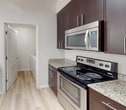 This efficiently designed 590-square-foot, one-bedroom, one-bathroom apartment offers a well-organized single-floor layout. Light hardwood-style flooring runs throughout the entire unit, contributing to a clean and cohesive aesthetic. The open-concept kitchen and living area serves as the heart of the home. The kitchen is equipped with stainless steel appliances — including an electric range, microwave, refrigerator, and dishwasher — complemented by dark espresso cabinetry, granite countertops, and a peninsula with pendant lighting. An in-unit washer and dryer are accessible directly from the kitchen. The living room receives natural light through large windows with views of surrounding greenery, and includes a wall-mounted HVAC unit. The bedroom measures 130 square feet and features a closet with built-in shelving, a window with blinds, and direct sightlines to the kitchen. The bathroom is appointed with a granite-topped vanity with an undermount sink, dark cabinetry, a tub/shower combo, and contemporary lighting. An exterior door with a full-lite window provides additional natural light to the unit.