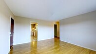 This single-floor residence offers a straightforward and functional layout designed for comfortable everyday living. The main living area is characterized by smooth hardwood flooring in a warm honey tone, which adds a welcoming brightness to the space. Clean white walls and a simple white ceiling contribute to an open, airy ambiance that makes the area feel well-lit and inviting. From the living area, an open doorway leads directly into the kitchen, which features wood-toned cabinetry and standard appliances, providing a practical and accessible cooking space. A second doorway off the living area opens to an additional room, finished with a deep green accent wall that provides a distinct visual contrast to the neutral tones of the main space. Baseboards run cleanly along the walls throughout, and the overall design maintains a consistent, understated aesthetic. The single-floor plan ensures easy navigation between the living, cooking, and adjoining spaces, making the layout efficient and cohesive. The space presents a clean canvas suitable for a variety of interior styling preferences.