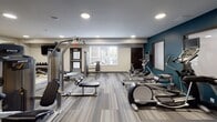 This fitness center is a comprehensively outfitted space designed to support a full range of workout routines. The room is divided into two distinct functional zones, each with its own flooring treatment. The strength training area features light wood-style flooring and is equipped with a multi-functional cable machine, a Smith machine with a weight bench, a dumbbell rack, and a flat bench. A large floor-to-ceiling mirror runs along one wall, allowing users to monitor their form during exercises.

The cardio zone transitions to a patterned, striped carpet tile floor and houses a row of treadmills and elliptical machines, each equipped with individual display screens. Additional cardio equipment, including rowing machines, is also present. A wall-mounted flat-screen television and a built-in dual water fountain with a bottle-filling station serve this area, adding practical convenience.

Throughout the space, recessed ceiling lights provide consistent, even illumination. A teal accent wall adds a bold visual contrast to the neutral grey and white walls. Natural light filters in through windows along one wall, and direct exterior access is available through a glass-paneled door. The overall design is functional and modern, well-suited for a variety of fitness needs.