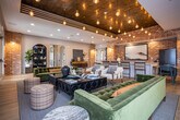 Located at 5060 City Station Dr in Collegedale, Tennessee, this single-floor community amenity space blends industrial and contemporary design into a cohesive, inviting environment. The open-plan layout is anchored by rich light hardwood flooring that flows throughout, unifying the various distinct zones within the space.

The lounge area features a tufted velvet sofa and curated seating arranged around a sleek linear fireplace, with a wall-mounted flat-screen TV above. French doors on either side flood the area with natural light and provide direct access to an outdoor patio. A striking coffered ceiling with warm wood paneling and cascading Edison bulb pendant lights adds architectural character and visual depth.

The dining and social zone showcases exposed brick accent walls that lend an industrial warmth to the space. A live-edge wood dining table paired with contemporary chairs serves as a focal point, complemented by a long white quartz bar with barstool seating. Additional pendant lighting in matte black with gold interiors illuminates a generous banquette upholstered in caramel leather against teal-painted walls.

Throughout the space, decorative brick columns, arched shelving displays, and curated artwork contribute to a layered, upscale aesthetic. The thoughtful combination of textures, materials, and lighting creates a versatile gathering space equally suited for casual socializing or structured group use.